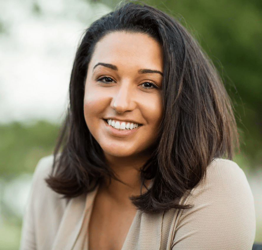 A woman with shoulder-length dark hair smiles warmly at the camera. She is wearing a beige top and is outdoors, with a blurred green background of trees, perfectly capturing the friendly vibe of the ScreenCoach Offer.