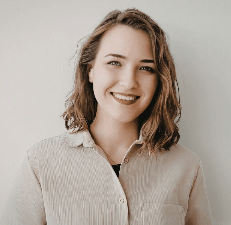 A young woman with wavy brown hair smiles at the camera. She is wearing a light-coloured, button-up shirt and stands against a plain, light background, ready to share her excitement about the latest ScreenCoach offer.