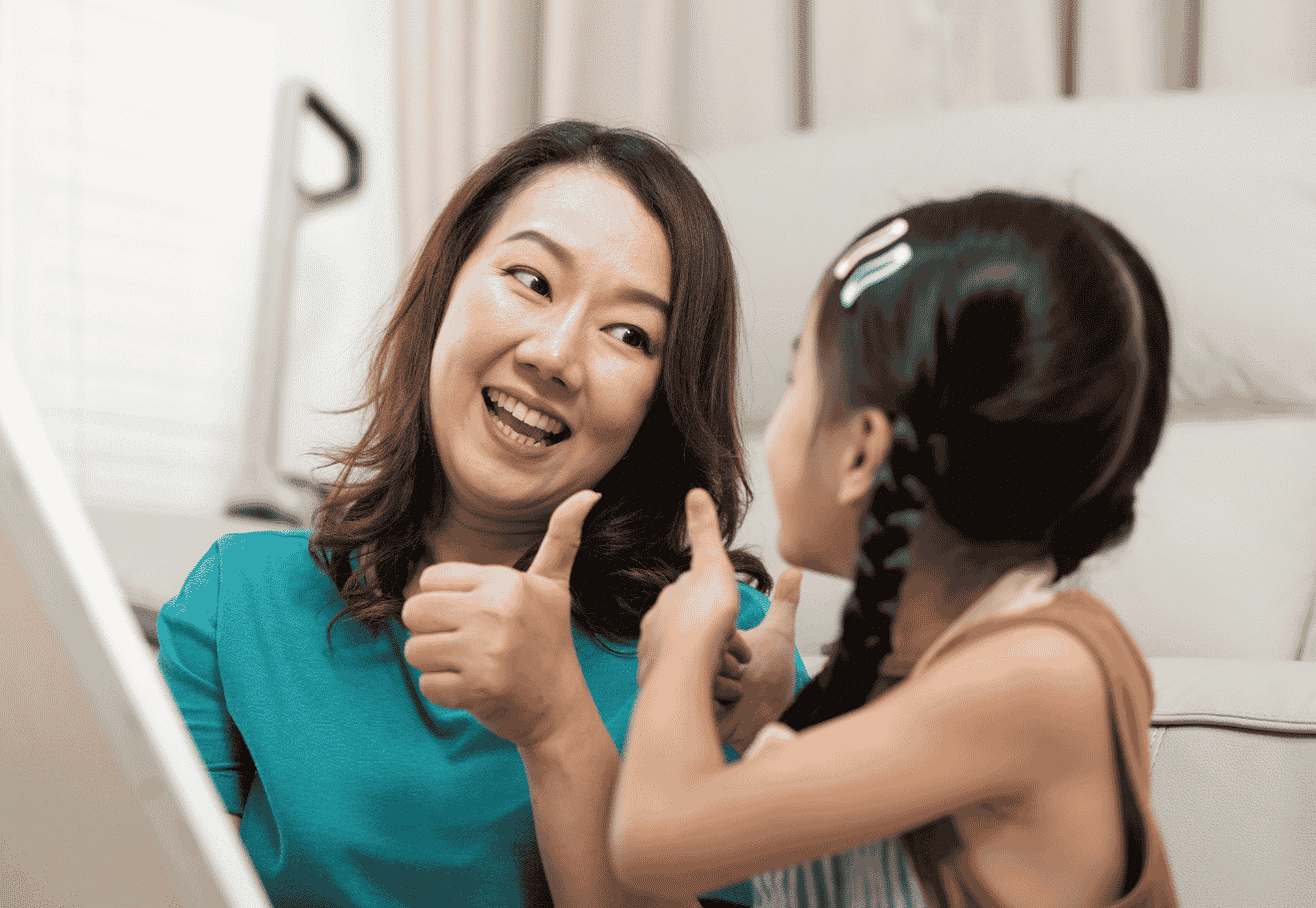 A woman and a young girl are smiling at each other, both giving a thumbs up. The woman has dark hair and wears a teal top; the girl has plaited hair with clips and faces the woman. They appear happy and engaged indoors, sharing a moment that could be celebrated using positive parenting apps to track rewards or manage screen time together.